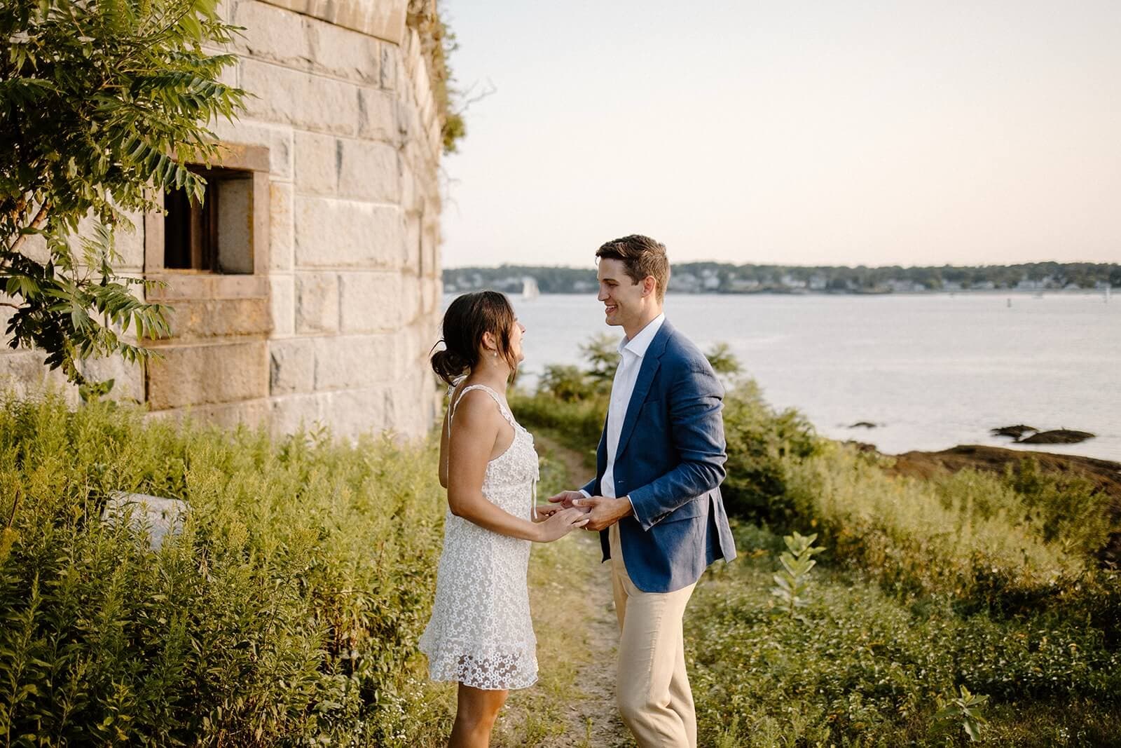 A couple holds hands and smiles at each other, standing on a grassy area by the water at sunset.